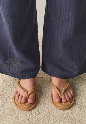 Fødder iført beige flettede sandaler og marineblå teksturerede bukser, der står på et lyst beige tæppe.