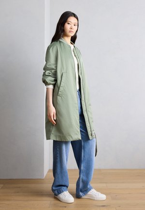 Frau steht drinnen und trägt eine lange, hellgrüne Bomberjacke, weiße Sneakers, hellblaue Jeans und ein weißes Oberteil.