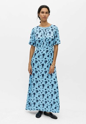 Femme vêtue d'une robe maxi à manches courtes bleu clair à motifs floraux, debout devant un fond blanc uni, chaussée de chaussures noires sans lacets.