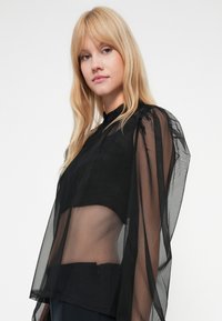 Top de malha preta com mangas longas e transparentes e um corpete ajustado. O design inclui um decote alto e um detalhe com zíper na bainha.