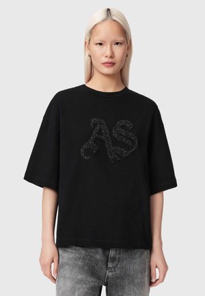 Jeune personne aux longs cheveux blonds portant un T-shirt noir surdimensionné avec un logo "AS" scintillant et un jean gris délavé, sur un fond uni.