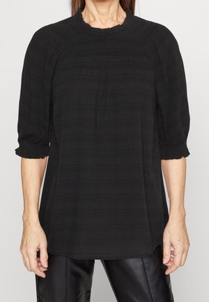 Blouse noire en tissu léger texturé, avec des manches courtes à volants et des bords festonnés, et un col rond.