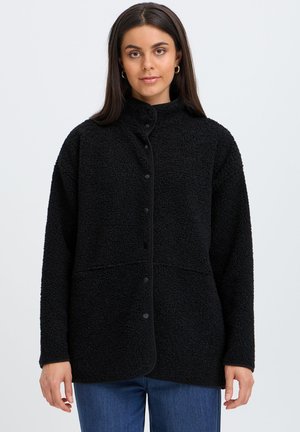Vrouw draagt een zwarte sherpa fleecejas met knopen en blauwe spijkerbroek, kijkt recht vooruit met een neutrale uitdrukking tegen een effen achtergrond.