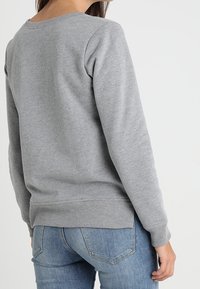 Mujer con una sudadera gris de manga larga y jeans azules, vista desde atrás contra un fondo blanco liso.