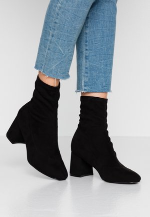 Bottes chevilles en daim noir avec design affaissé, bout pointu et talon épais. Portées avec un jean court bleu clair. Texture lisse.