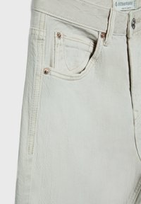 Jeans en denim couleur crème présentant un design classique, des coutures ton sur ton, une poche avant avec un rivet en cuivre et une finition en tissu texturé.