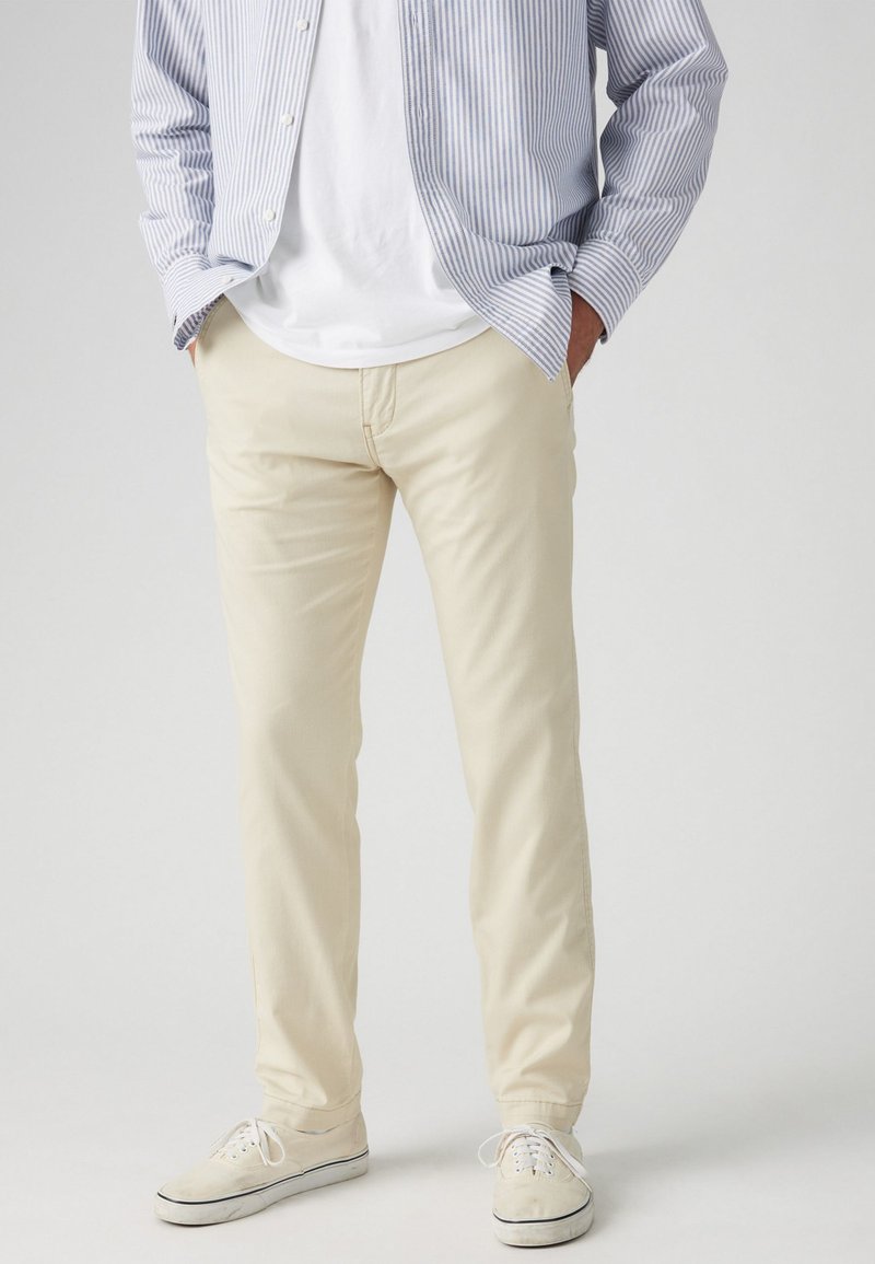 Pantalons en coton beige clair avec une coupe droite, texture subtile et poches standard, associés à des baskets blanches et une chemise rayée bleue.