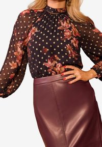 Svart blommig blus med goldiga prickar, veckad halsringning och genomskinliga långärmar, ihop med en figurnära bordeaux läderkjol.