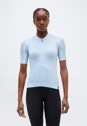 Femme debout, de face, portant un maillot de cyclisme à manches courtes bleu clair avec zip et un short de cyclisme noir.