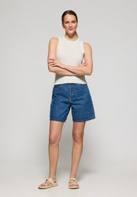 Shorts in denim blu medio con vestibilità rilassata, dotati di chiusura con bottone anteriore e tasche. Abbinati a un top senza maniche color beige chiaro.
