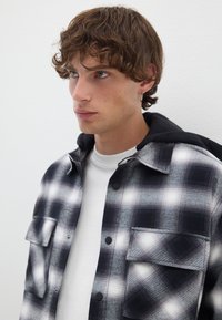 Schwarz-weißer kariert Flanellhemd mit zwei vorderen Taschen, getragen über einem weißen T-Shirt, mit einer schwarzen Kapuze und strukturiertem Stoff.