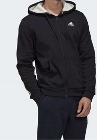 Sweat à capuche noir zippé avec doublure polaire blanche dans la capuche, logo Adidas sur la poitrine, porté par une personne avec les mains détendues sur les côtés.