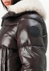 Vrouw in een glanzende donkerbruine pufferjack met een witte fleece kraag en een ronde zwarte "Save the Duck" logo op de bovenarm.