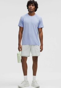 Jasnofioletowa koszulka sportowa z krótkim rękawem o subtelnej teksturze, w połączeniu z jasnoszarymi szortami i białymi sneakersami. Trzyma zieloną butelkę na wodę.