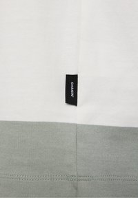 Weißer und hellgrauer Stoff mit einem kleinen schwarzen rechteckigen Etikett, auf dem "CHASIN'" steht, das in die Naht genäht ist.