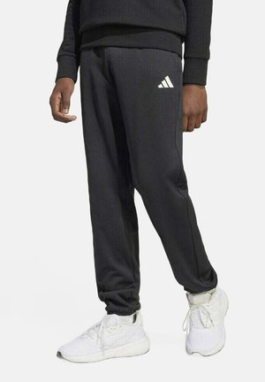 Pantalons de survêtement noirs Adidas avec taille élastique, jambes fuselées et logo blanc sur la hanche gauche, associés à des baskets de sport blanches.