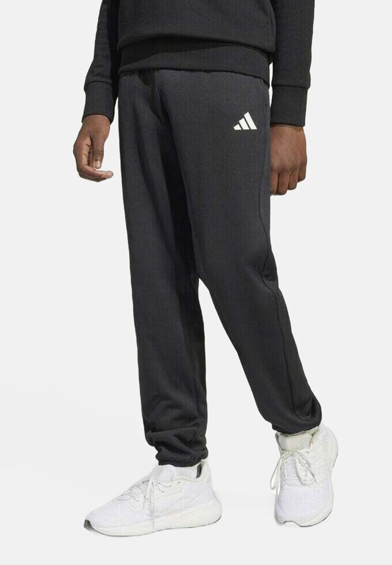 Zwarte Adidas sweatpants met een elastische tailleband, taps toelopende pijpen en een wit logo op de linkerheup, gecombineerd met witte sport sneakers.
