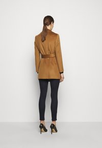 Mujer de espaldas vestida con un abrigo marrón con cinturón, leggings negros y zapatos de tacón negro, de pie contra un fondo blanco liso.