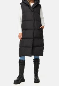 Schwarze Steppweste mit Kapuze, mit durchgestepptem Design, einem Frontverschluss in der Mitte und seitlichen Taschen, getragen über einem weißen Pullover und blauen Jeans.