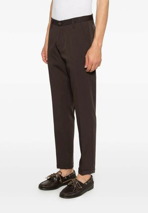 Uomo che indossa pantaloni marrone scuro su misura e mocassini in pelle marrone scuro, in piedi davanti a uno sfondo bianco.