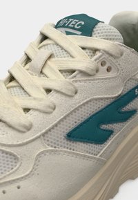 Λευκό αθλητικό παπούτσι με υφές από δίχτυ και σουέτ, με τόνους τιρκουάζ και παχιά μπεζ σόλα. Οι κορδόνια είναι ανοιχτό μπεζ και περιλαμβάνουν τρύπες.