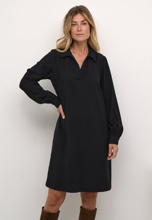 Schwarzes Kleid mit einem kragenlosen Ausschnitt, langen Puffärmeln und lockerer Passform. Besitzt eine weiche Textur und ein dezentes Detail an der Vorderseite.