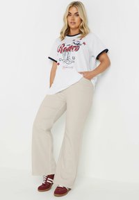 Vit grafisk t-shirt med blå detaljer som visar cowboyboots och stjärnor, kombinerad med beiga utsvängda byxor och röda sneakers.