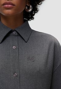 Camicia grigia con colletto, in un tessuto testurizzato, con un logo ricamato sottile e bottoni di un leggero colore viola. Orecchini a cerchio rotondi in argento.