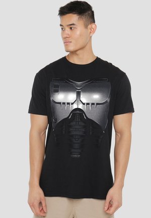 Man wearing black t-shirt with a shiny silver futuristic mask design on front, beige pants, looking to his left against a plain gray background.