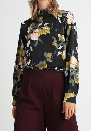 Frau trägt eine schwarze, langärmelige Bluse mit gelben, rosa und grünen Blumen, kombiniert mit hoch taillierter dunkelburgunder Hose.
