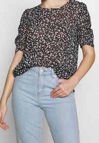 Blouse noire transparente à imprimé floral blanc, col rond et manches courtes bouffantes, associée à un jean bleu clair à taille haute.