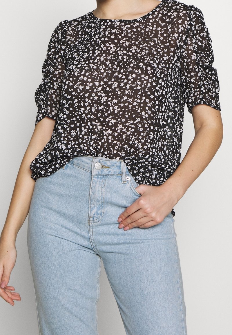 Blouse noire transparente à imprimé floral blanc, col rond et manches courtes bouffantes, associée à un jean bleu clair à taille haute.