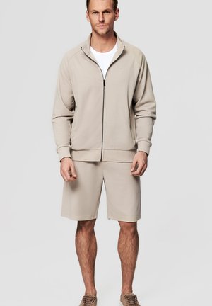 Mężczyzna ubrany w beżową kurtkę na zamek błyskawiczny na białą koszulę, do tego beżowe krótkie spodenki i beżowe trampki, stojący na jednolitym jasnym tle.