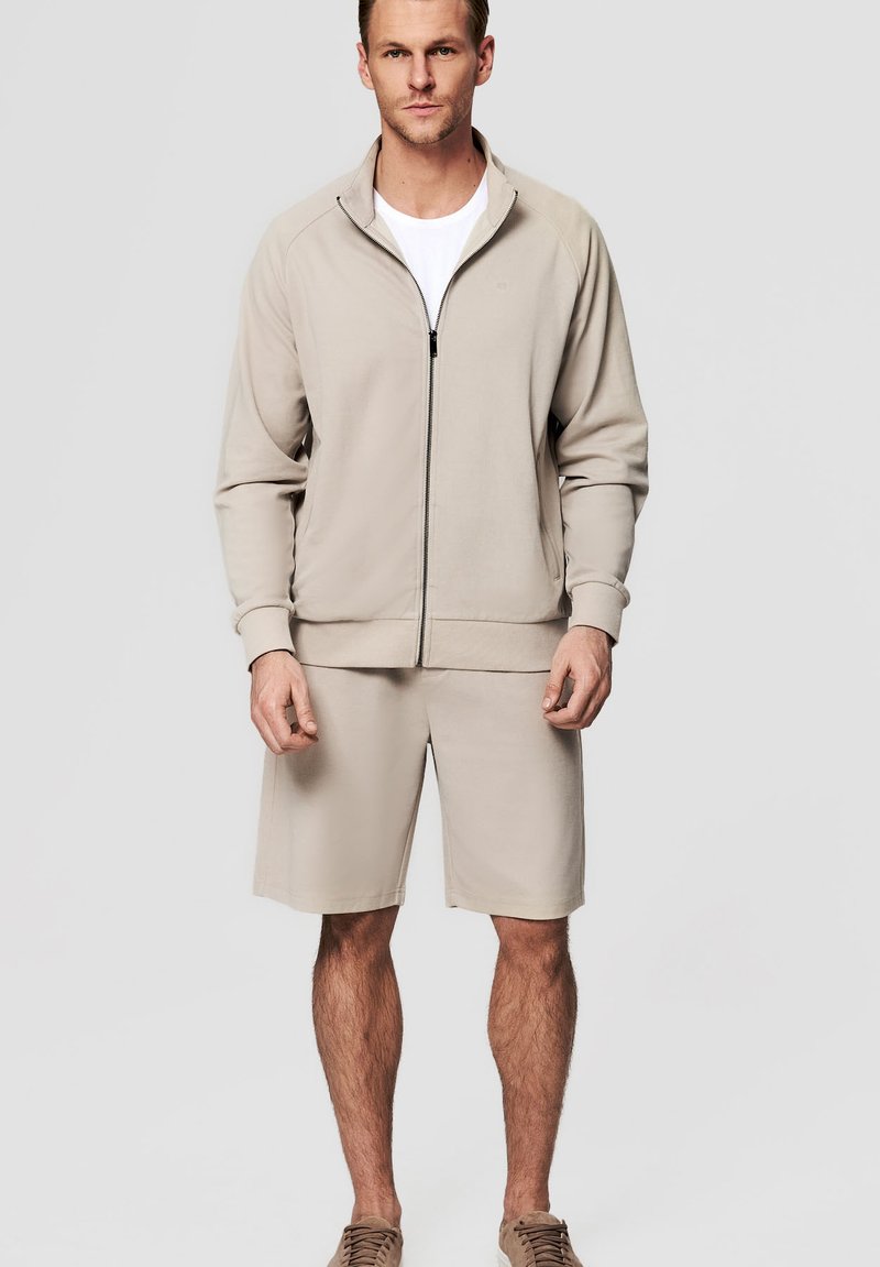 Mężczyzna ubrany w beżową kurtkę na zamek błyskawiczny na białą koszulę, do tego beżowe krótkie spodenki i beżowe trampki, stojący na jednolitym jasnym tle.