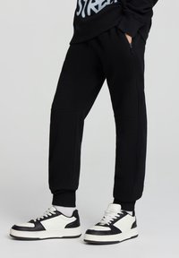 Czarne dresy z elastycznymi mankietami, z kieszenią na zamek. Połączone z czarno-białymi trampkami z teksturowanym wykończeniem.