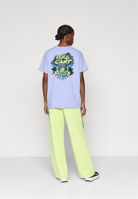 Λιλά μπλουζάκι με πολύχρωμο γραφικό σχέδιο στην πλάτη, συνδυασμένο με ψηλόμεσα, φαρδιά παντελόνια σε έντονο λουιζάτη πράσινη απόχρωση.