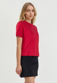Jonge vrouw met blond haar, gekleed in een rode T-shirt met korte mouwen en een zwarte rok, staande tegen een effen witte achtergrond.