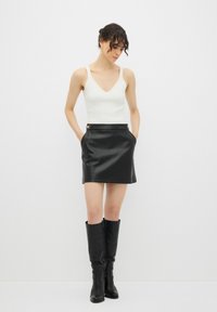 Zwarte leren mini-jurk met zijzakken en gouden accenten, gecombineerd met een aangesloten witte tanktop en kniehoge zwarte laarzen.