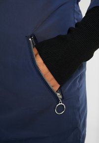 Granatowa odzież wierzchnia z kieszenią na zamek. Posiada teksturowany czarny mankiet i srebrne okucia zamka zapewniające bezpieczne zamknięcie. Gładka, wodoodporna tkanina.