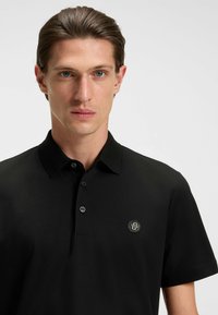 Czarna koszulka polo wykonana z gładkiej bawełny, z klasycznym kołnierzykiem, zapięciem na trzy guziki oraz naszywką z logo na lewej piersi.