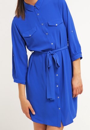 Vrouw draagt een koningsblauwe, knielange jurk met lange mouwen, knopen en borstzakken, en een ceintuur in de taille die is gestrikt.