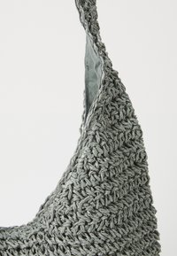 Gros plan d'un sac à bandoulière gris en crochet montrant la texture tissée détaillée et la doublure en tissu sur un fond uni.