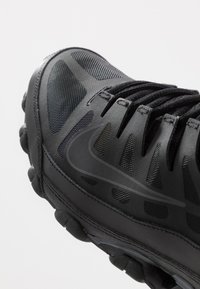Black athletic shoe with a textured mesh upper, accented logo, cushioned sole, and thick laces, designed for performance and comfort.