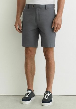 Mężczyzna ubrany w szare, dopasowane szorty, białą koszulę i czarne trampki z białymi sznurówkami, stojący na tle jednolitej białej ściany.