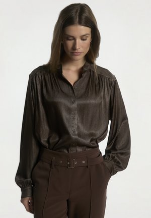 Vrouw met steil bruin haar draagt een glanzende bruine blouse met knopen, ingestopt in een hoge taille bruine broek met een brede riem, handen in de zakken.