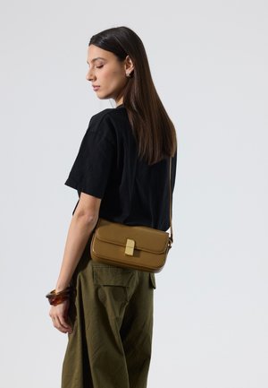 Vrouw met steil bruin haar draagt een zwart T-shirt, olijfgroene broek, barnstenen armband en draagt een bruine leren schoudertas met gouden gesp.