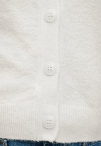 Κοντινό σε λευκό πλεκτό ζακέτα με τρία λευκά κουμπιά και ριμπ τελείωμα πάνω από μπλε τζιν παντελόνι.