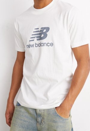 Osoba nosząca białą koszulkę New Balance z szarym logo i napisem, w połączeniu z jasnoniebieskimi jeansami, z jedną ręką w kieszeni.