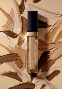Estée Lauder Futurist concealer i en genomskinlig tub med svart lock, omgiven av prover av olika hudtoner på en beige bakgrund.