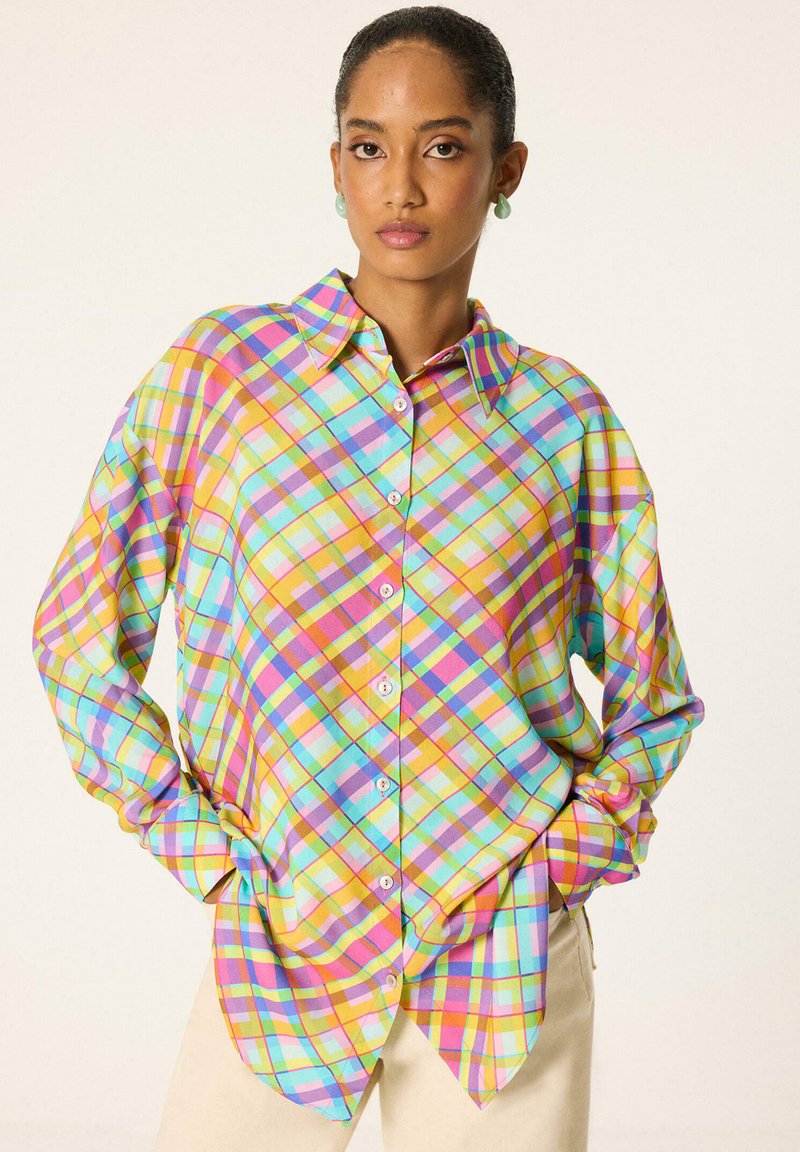 Femme portant une chemise à carreaux pastel colorée à manches longues, debout les mains dans les poches devant un fond uni.